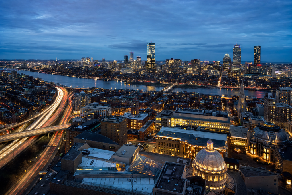 High-vantage view of Boston during blue hour, glowing city lights, subtle motion blur in traffic