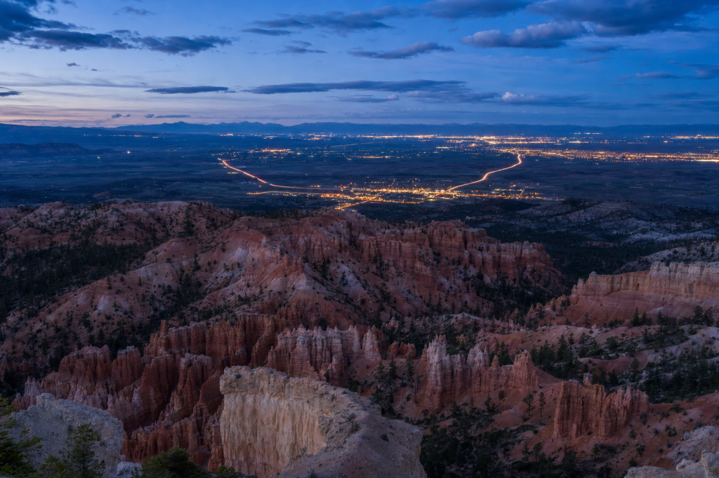 High-vantage view of Bryce Canyon during blue hour, glowing city lights, subtle motion blur in traffic
