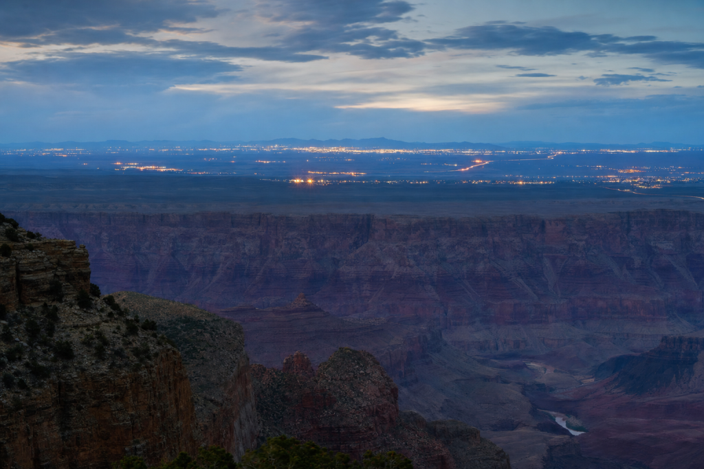 Atmospheric high-vantage view of Grand Canyon during blue hour, glowing city lights, subtle motion blur in traffic