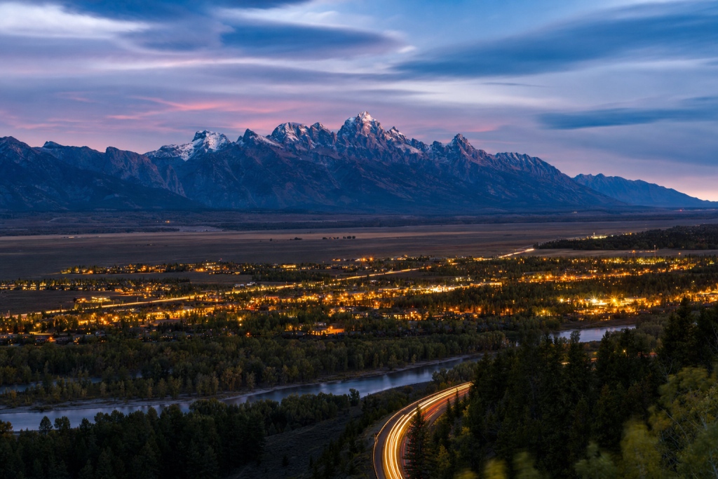 High-vantage view of Grand Teton during blue hour, glowing city lights, subtle motion blur in traffic
