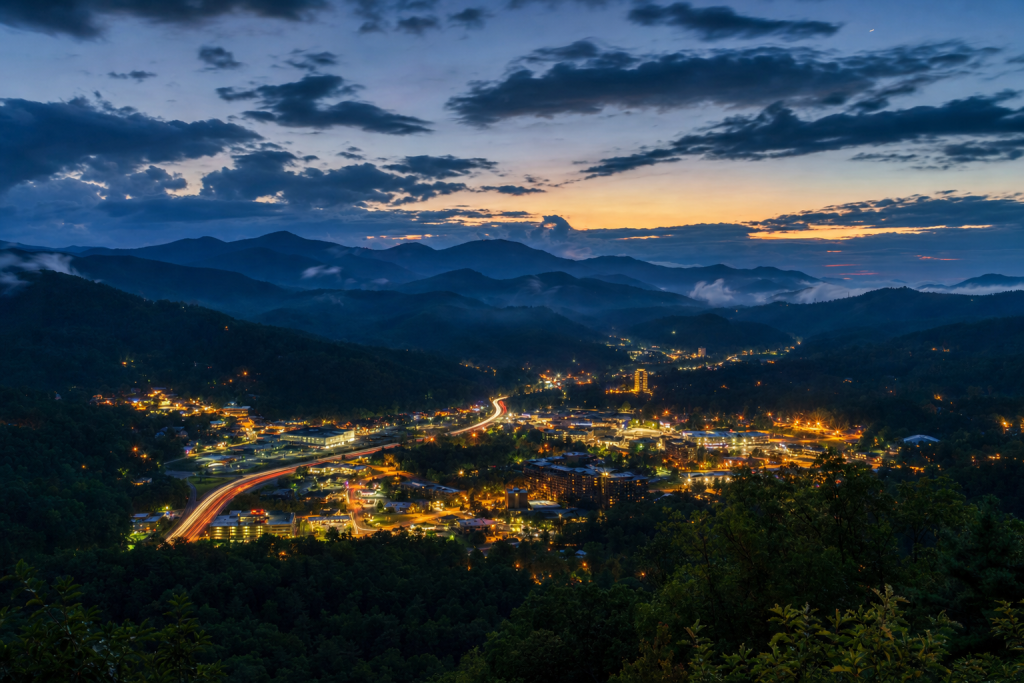 Atmospheric high-vantage view of Great Smoky Mountains during blue hour, glowing city lights, subtle motion blur in traffic