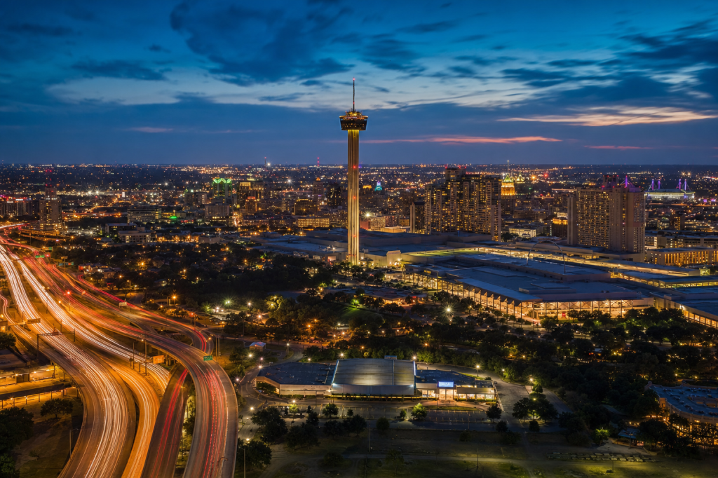 Atmospheric high-vantage view of San Antonio during blue hour, glowing city lights, subtle motion blur in traffic