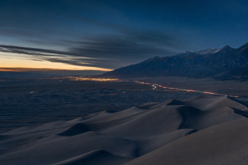 High-vantage view of Great Sand Dunes during blue hour, glowing city lights, subtle motion blur in traffic