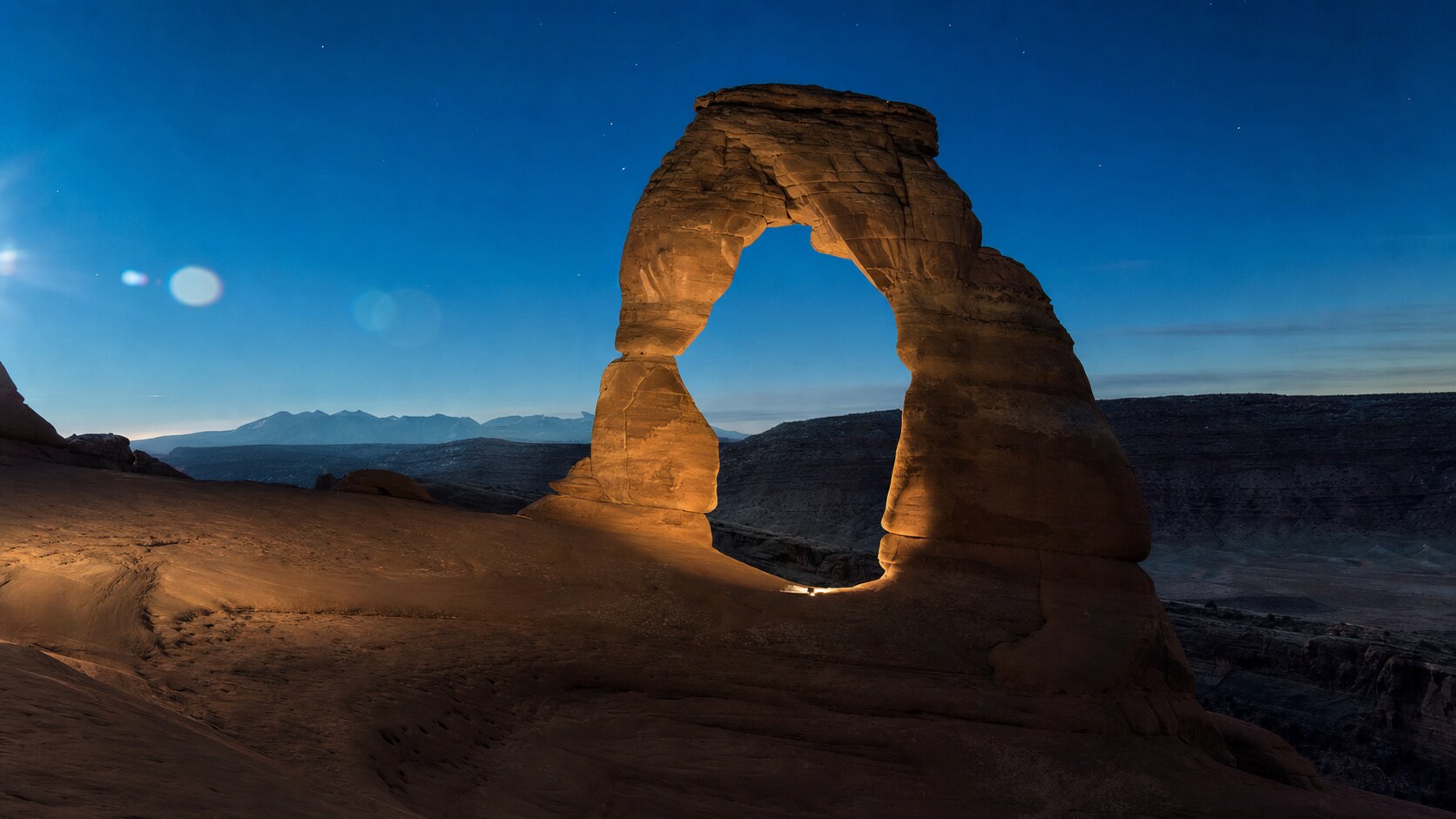 Wide blue-hour view of Delicate Arch with cobalt sky and warm artificial lighting on the landmark