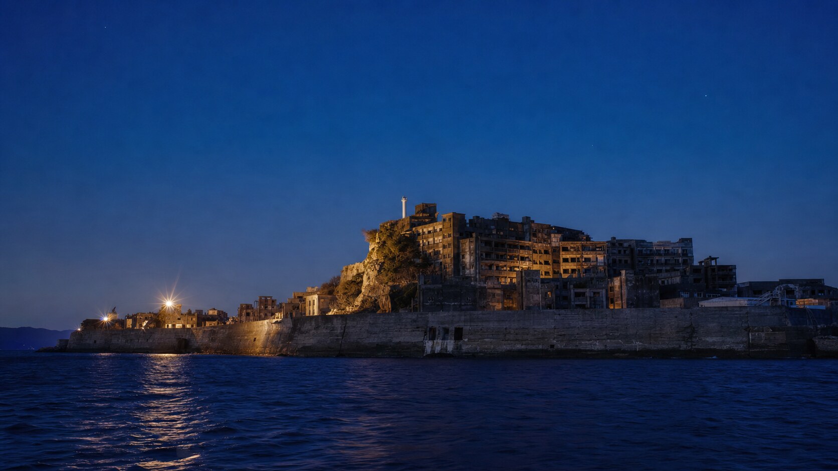 Wide blue-hour view of Hashima Battleship Island with cobalt sky and warm artificial lighting on the landmark