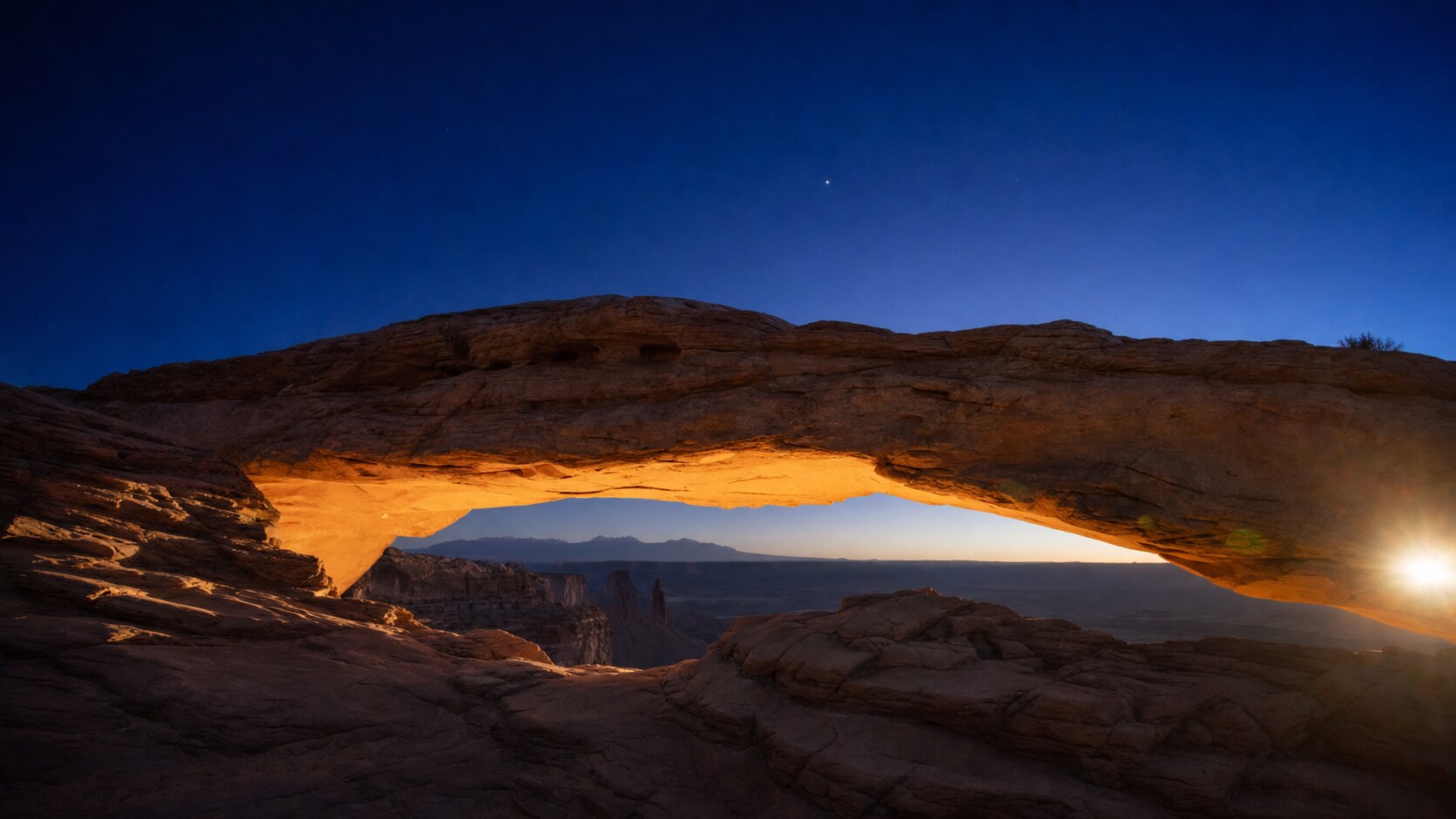 Wide blue-hour view of Mesa Arch with cobalt sky and warm artificial lighting on the landmark