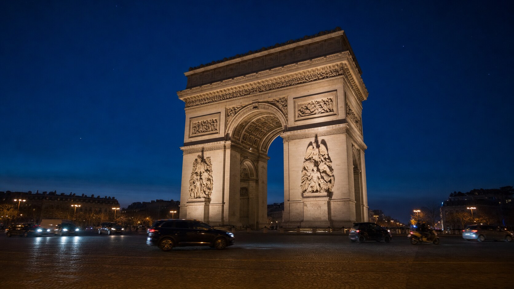 Wide blue-hour view of The Arc De Triomphe with cobalt sky and warm artificial lighting on the landmark