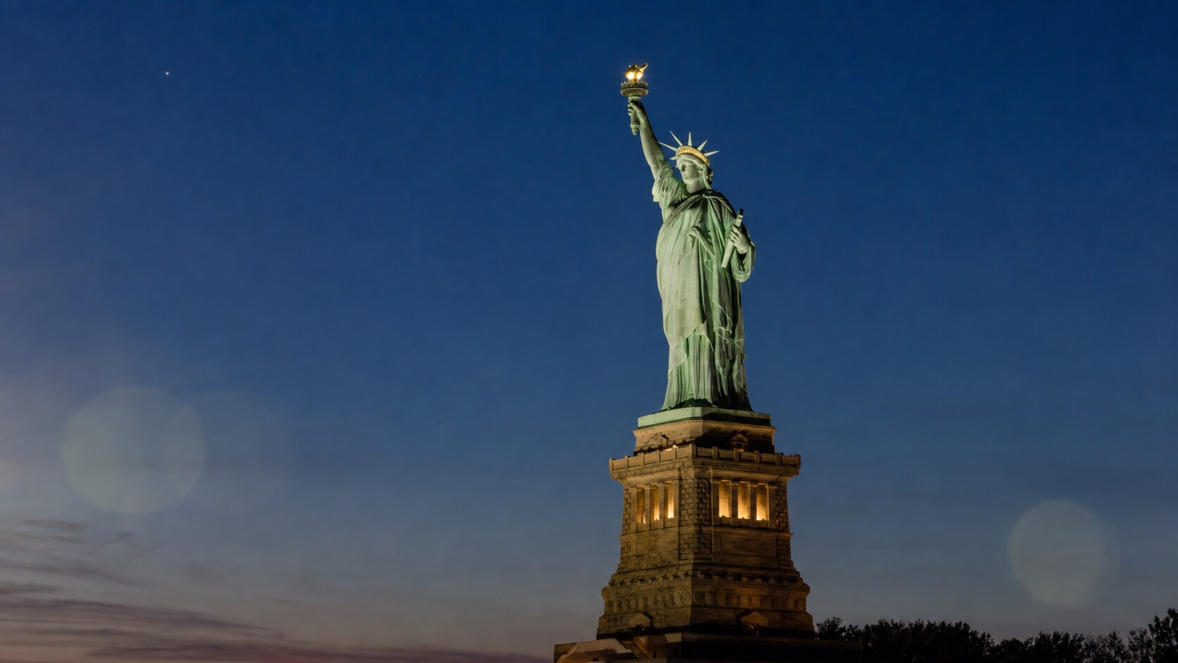 Wide blue-hour view of The Statue Of Liberty with cobalt sky and warm artificial lighting on the landmark