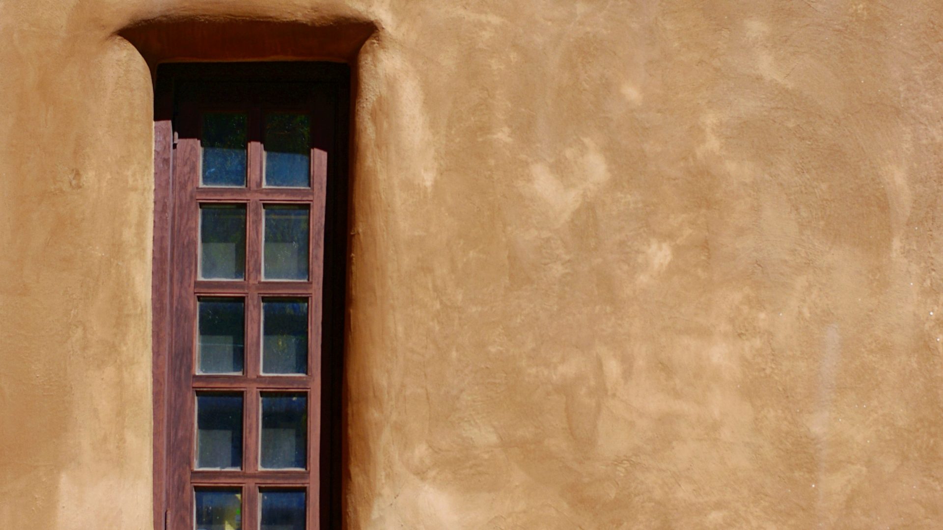 Wood-framed window and deep beam shadows on a warm adobe wall in Santa Fe, New Mexico.