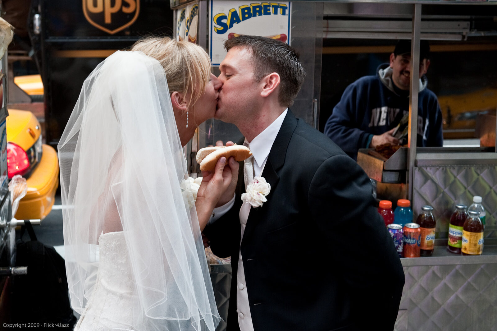 Bride and groom sharing a kiss at a Times Square hot dog stand, yellow taxis and city crowds blurred behind them