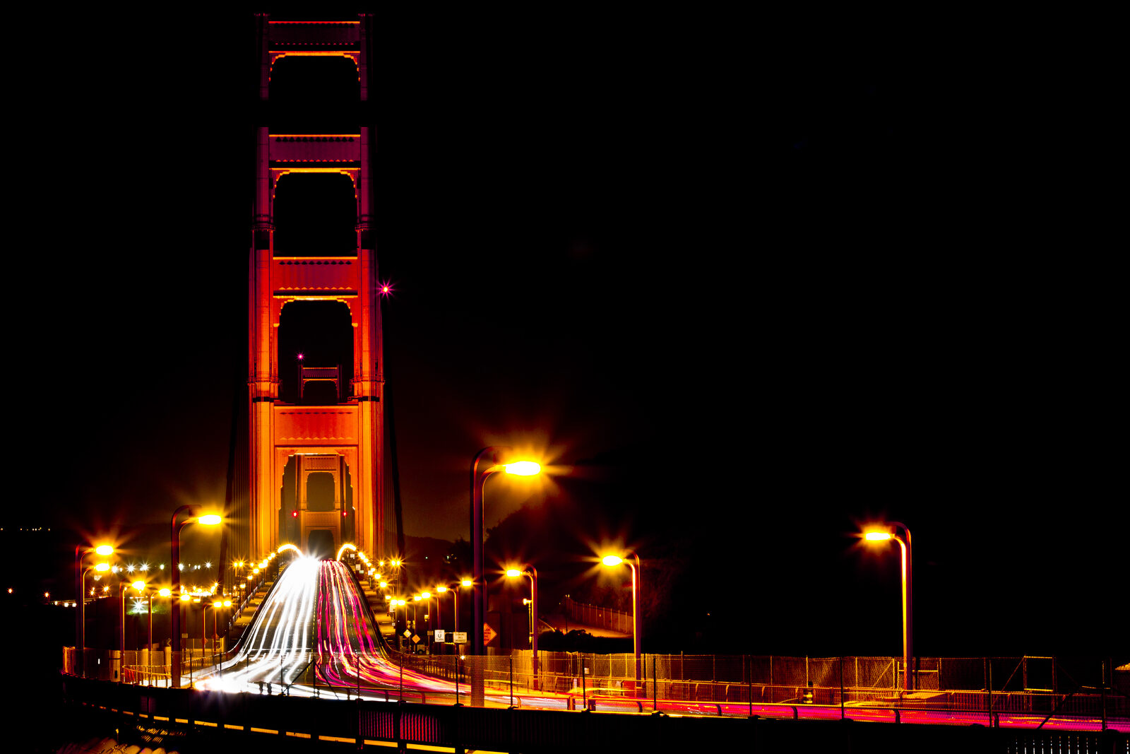 Golden Gate Bridge at night with red car light trails streaking across the deck and dark San Francisco bay