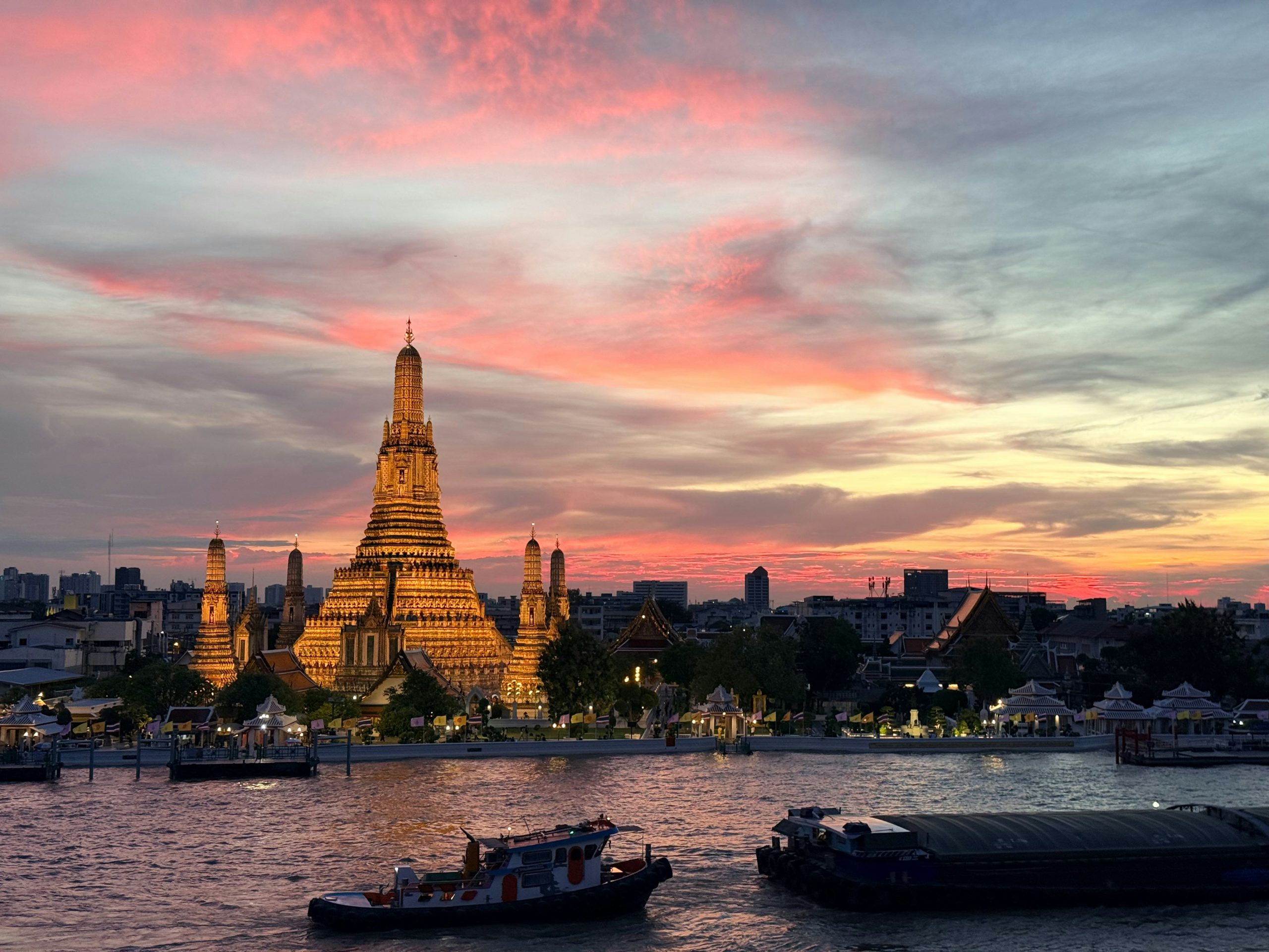 Illuminated Wat Arun across the Chao Phraya River beneath a pink sunset sky, with boats in the foreground