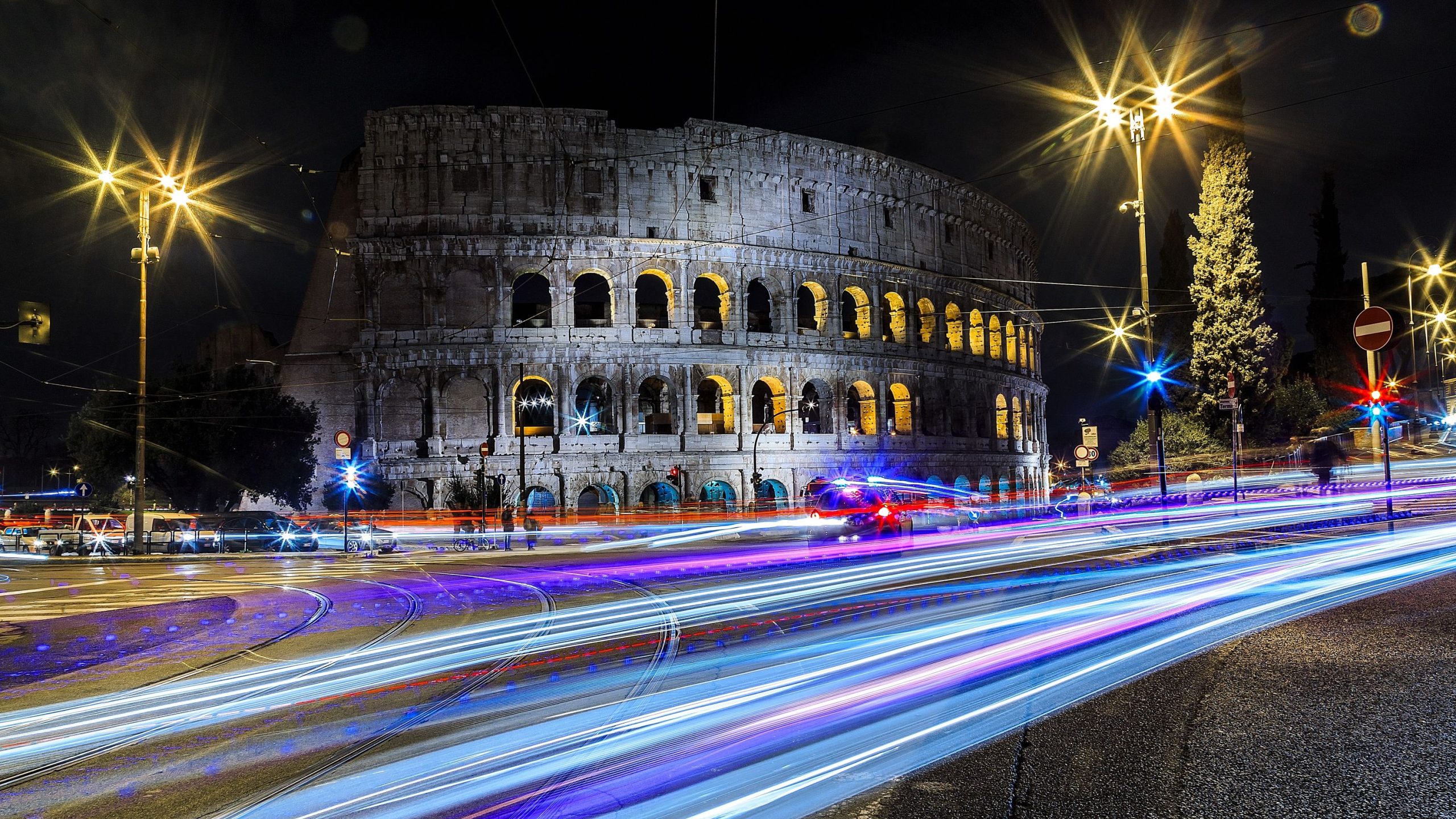 Colosseum in Rome illuminated at night, with colorful traffic light trails crossing the foreground