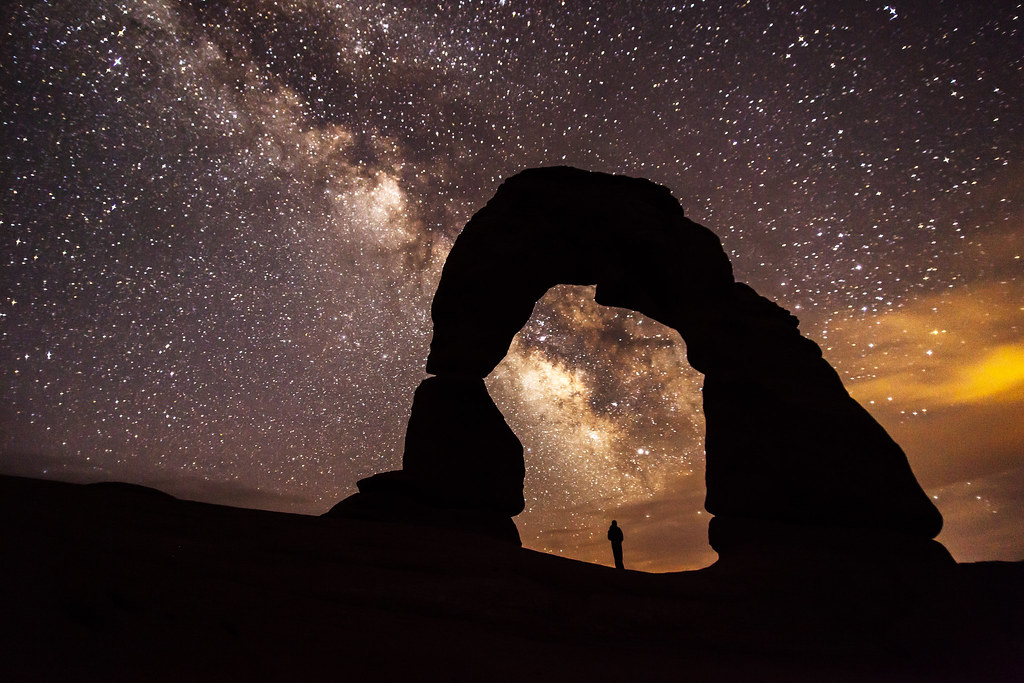 Delicate Arch silhouetted beneath the Milky Way, with a person standing beneath it at night