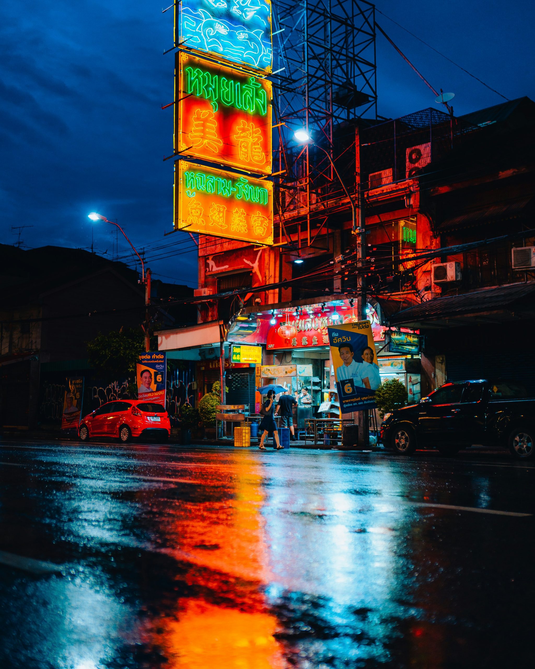 Neon signs above a wet street in Bangkok's Chinatown at blue hour, with colorful reflections on the road.