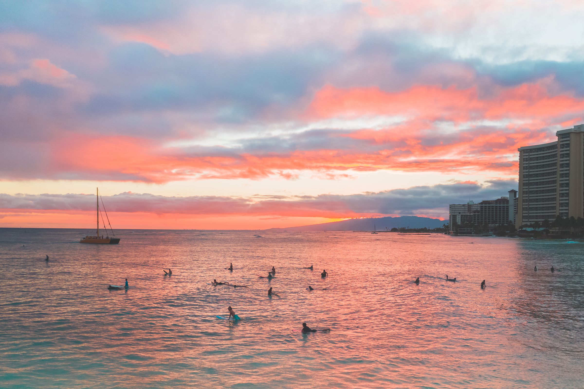 Surfers in the water off Waikiki Beach as pink sunset clouds fill the Honolulu sky