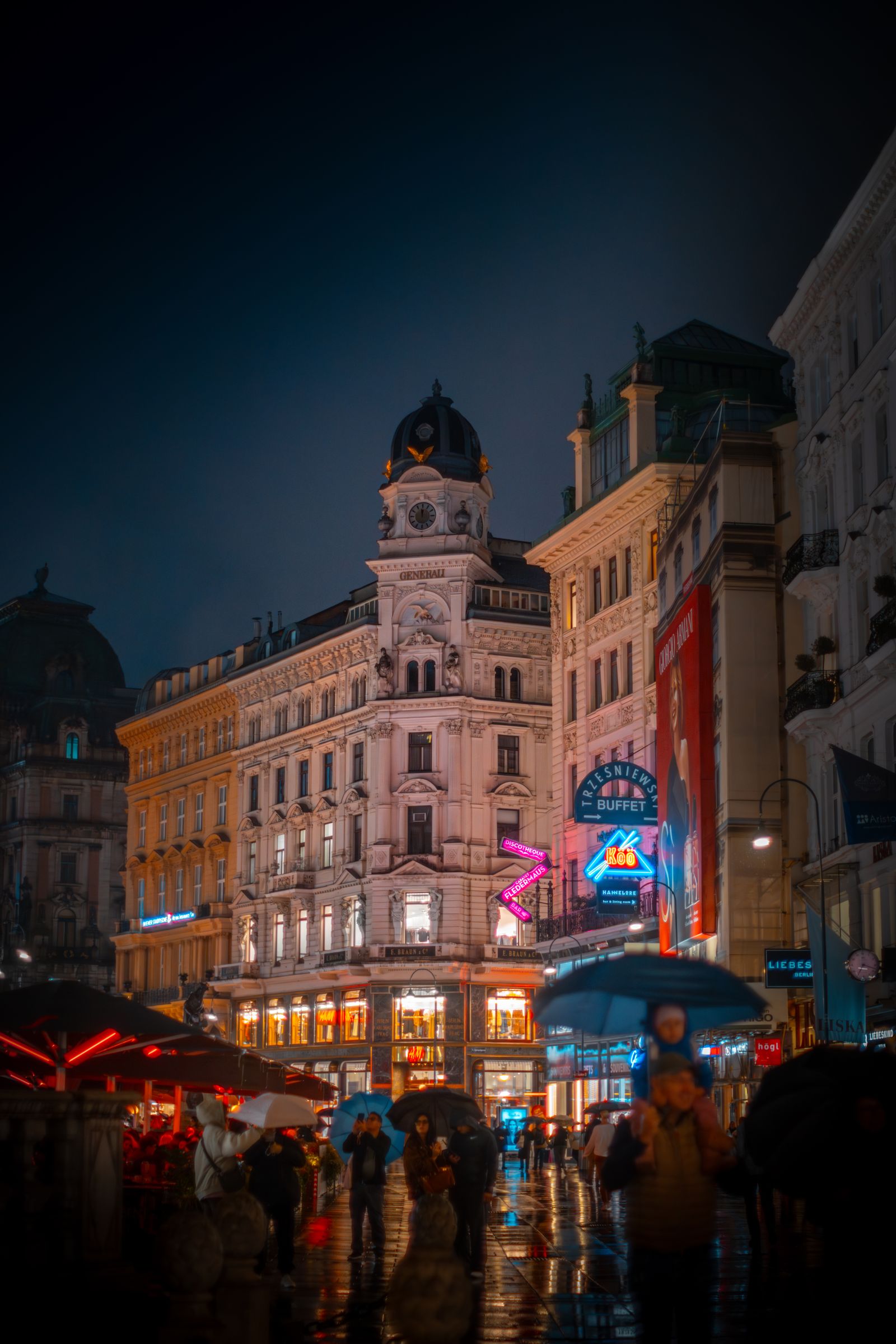 People under umbrellas on a wet Vienna street at night beneath illuminated historic buildings