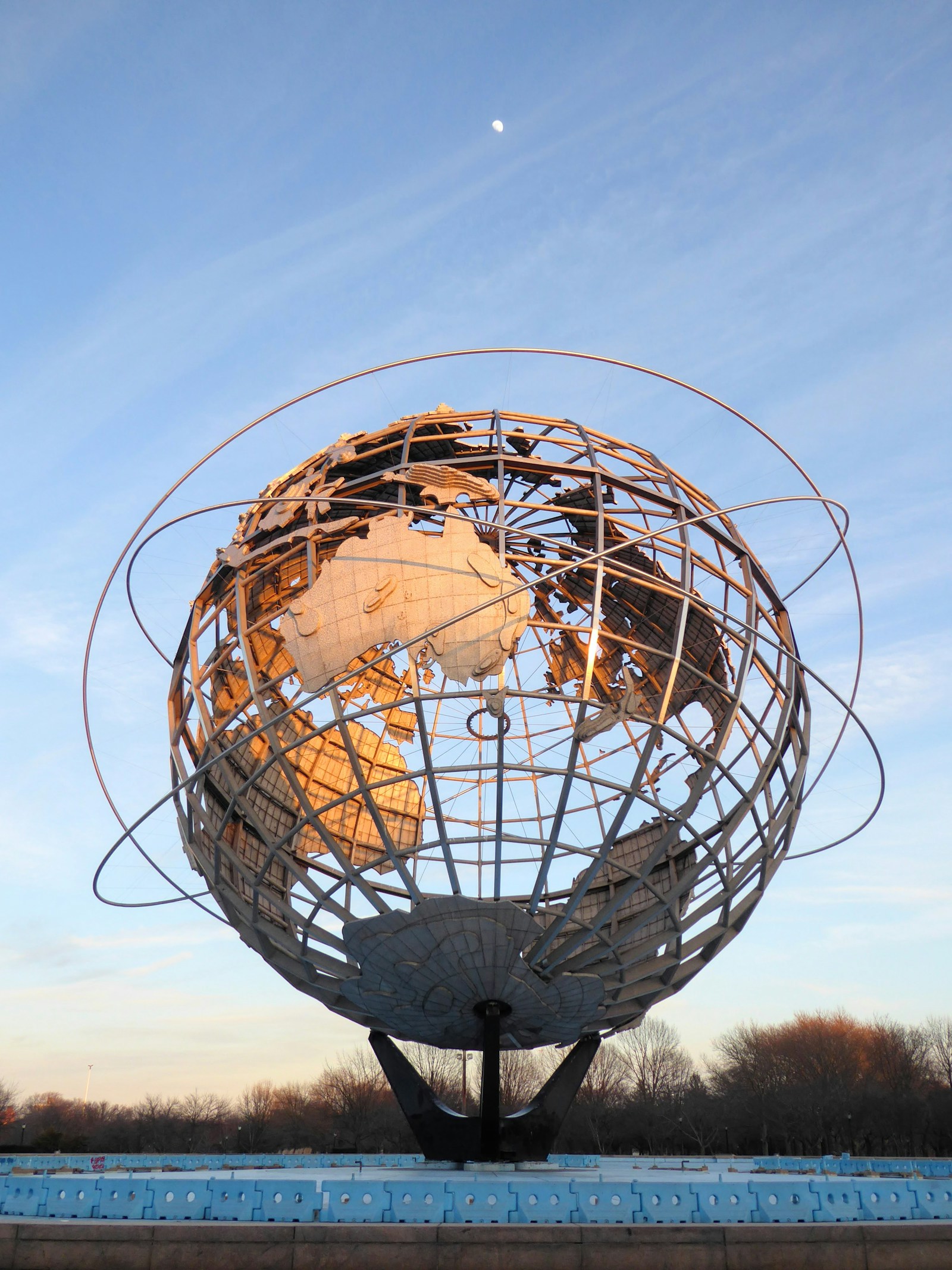 Unisphere globe lit by low sun beneath a blue sky and small moon