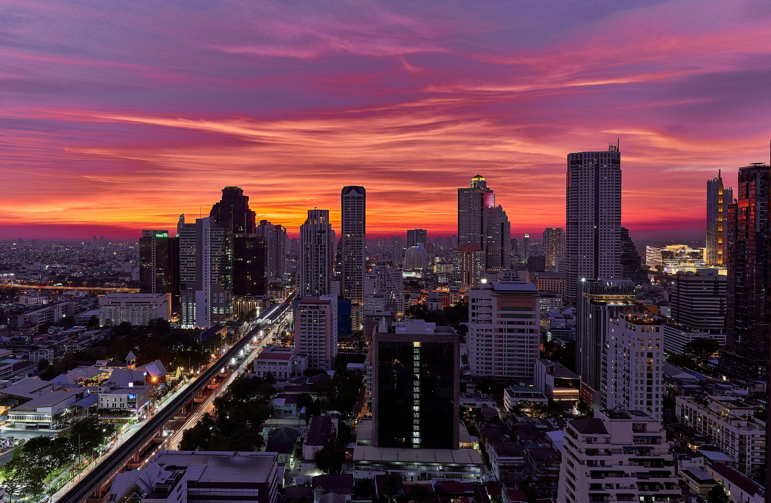 Bangkok skyline and elevated railway below a magenta-orange sunset sky