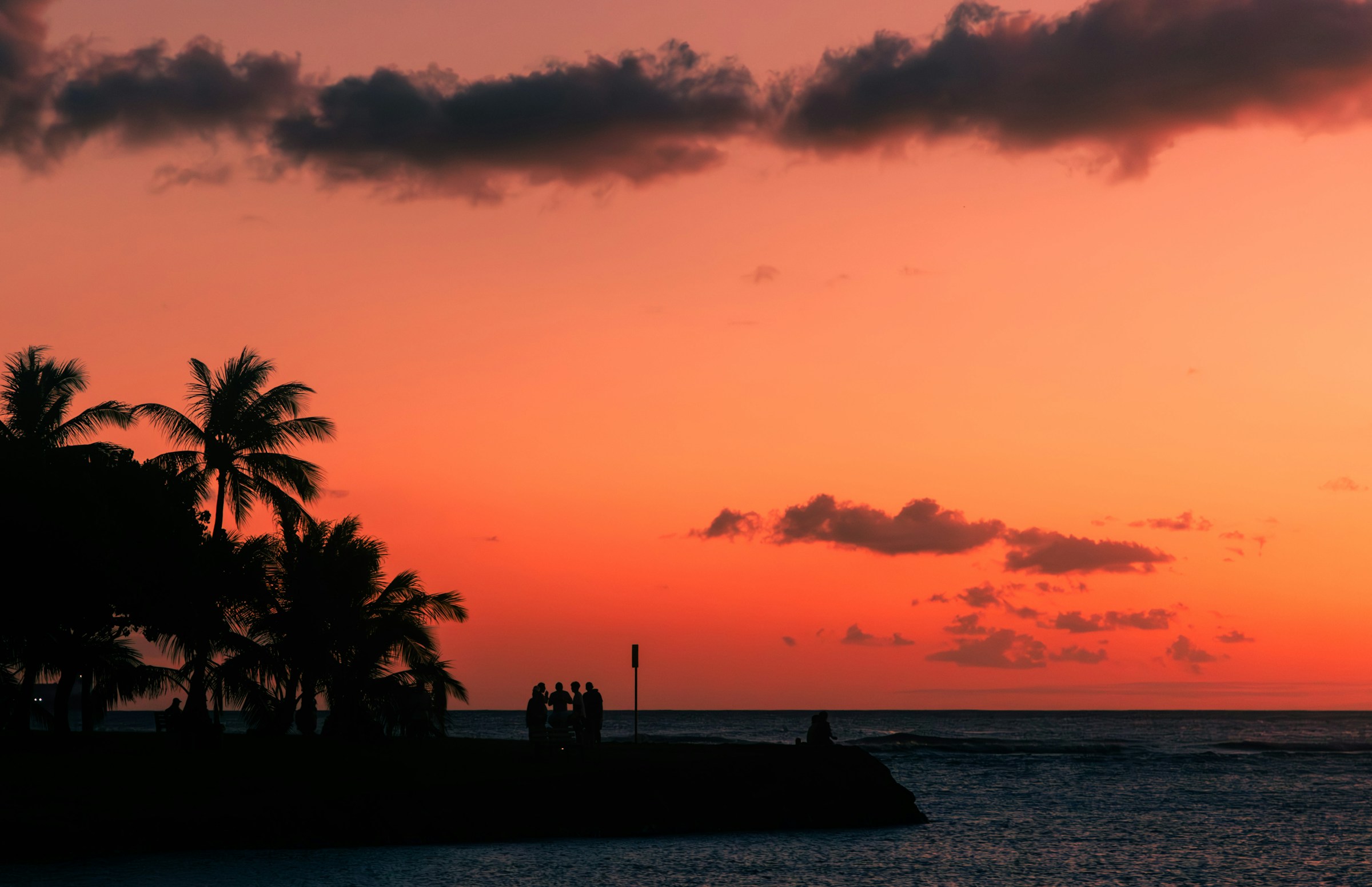 Palm trees and people silhouetted against coral sunset at Ala Moana Beach Park, Honolulu