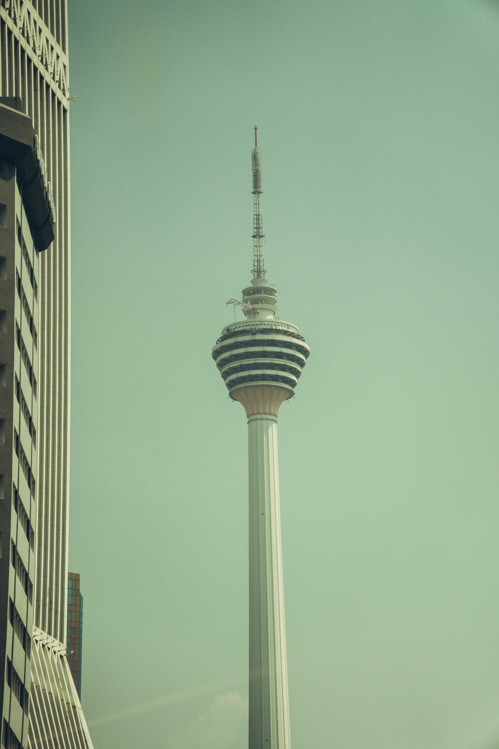 KL Tower rising beside a high-rise building under a pale sky.