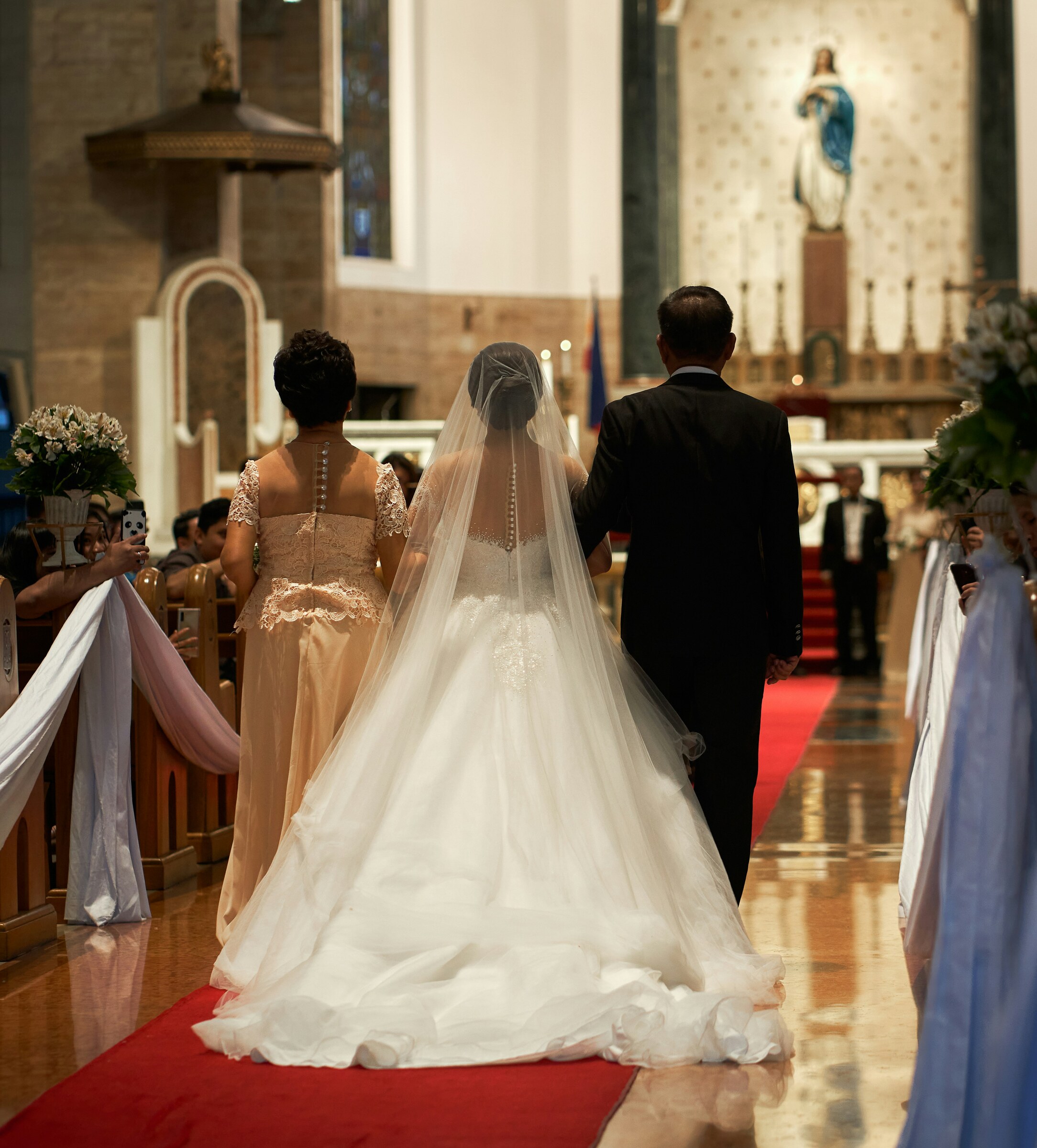 Bride in a long veil walking down a church aisle with her father and another companion toward the altar.