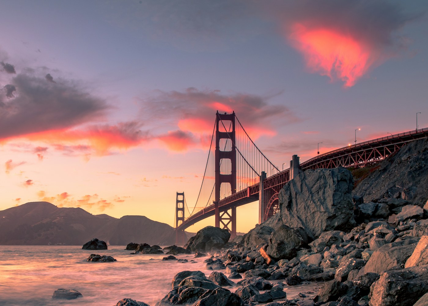 Golden Gate Bridge beneath vivid pink and orange clouds at sunset from the rocky shore