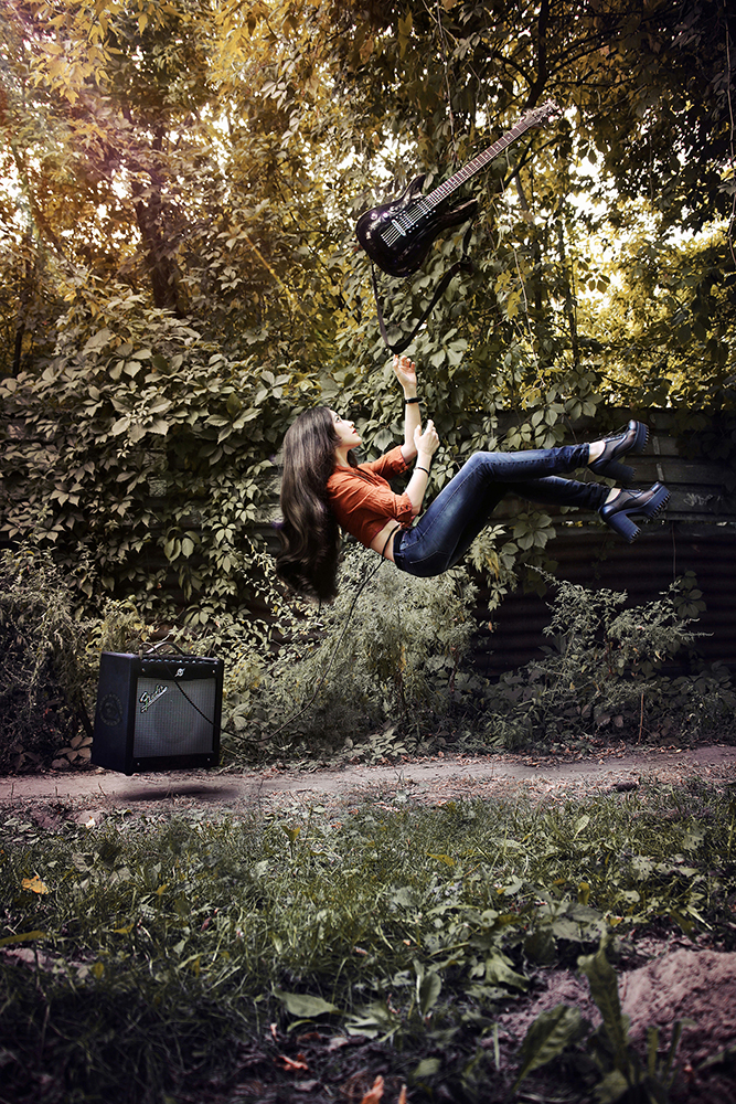 Person holding a guitar appears suspended sideways in a wooded clearing.