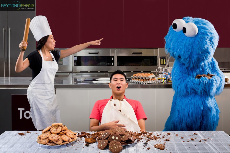 Bride in a chef hat gestures across a kitchen while a groom sits beside a blue mascot.