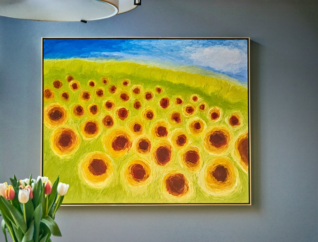 Framed painting of yellow and orange sunflowers on a gray wall, with tulips at lower left.