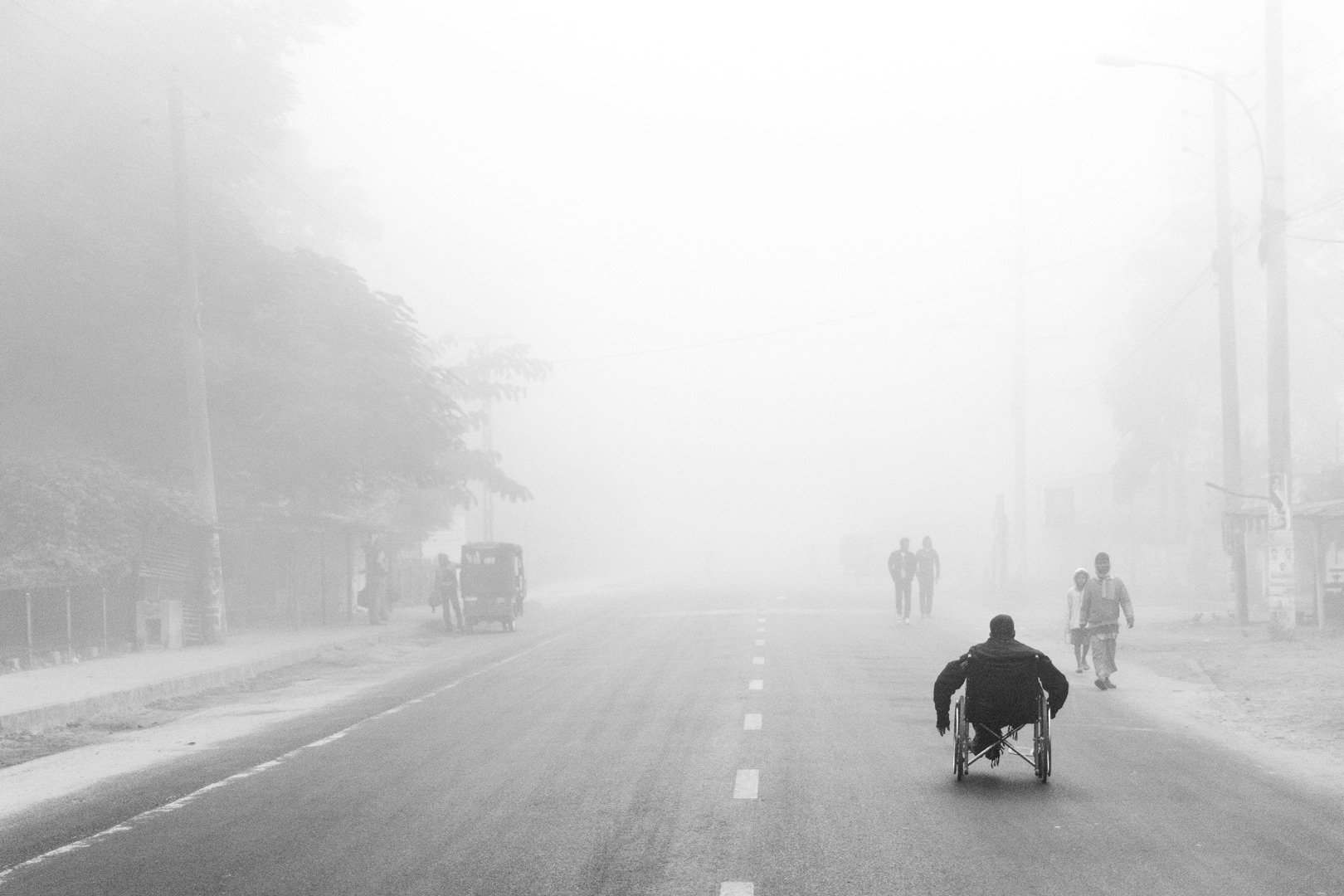 A man in a wheelchair on a broad road with pedestrians in heavy fog.