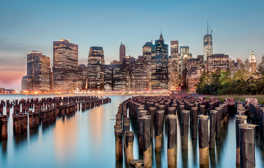 Lower Manhattan skyline seen beyond wooden pilings by the waterfront.
