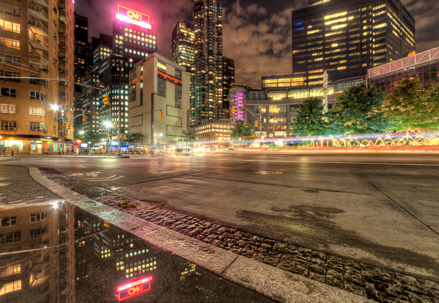 Lit Manhattan street at night with traffic light trails and puddle reflections.