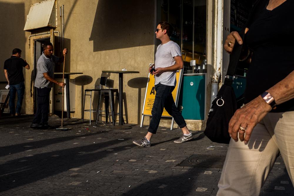 People pass a small take-away restaurant on a sunlit sidewalk.