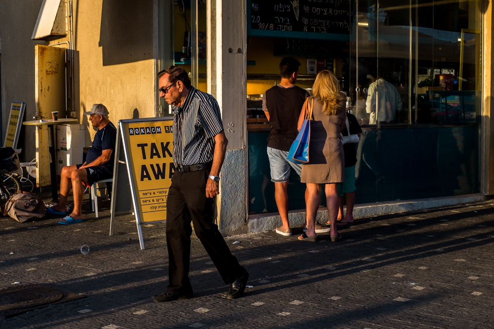 Man walks past a yellow take-away sign while people stand at a shop window.
