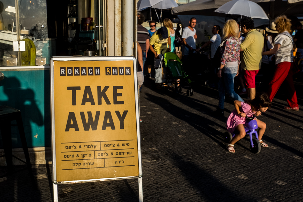 A take-away sign stands on a sidewalk beside a crowd and a small child.