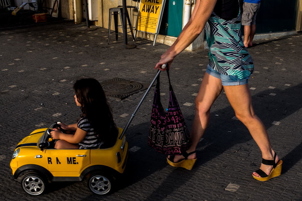A child rides in a small yellow toy car as pedestrians pass on the sidewalk.