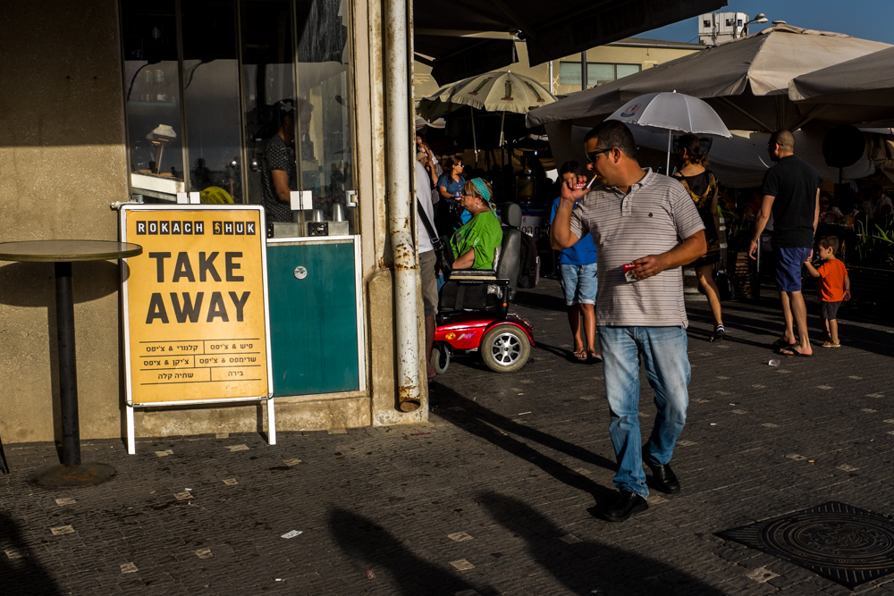 Man drinking from a cup walks past a take-away sign on a sunny sidewalk.