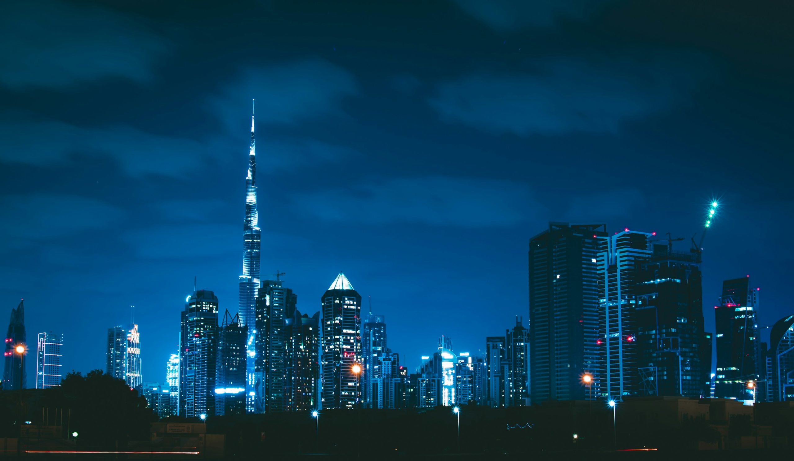 Dubai skyline at blue hour, with the illuminated Burj Khalifa rising above buildings under dark blue clouds.