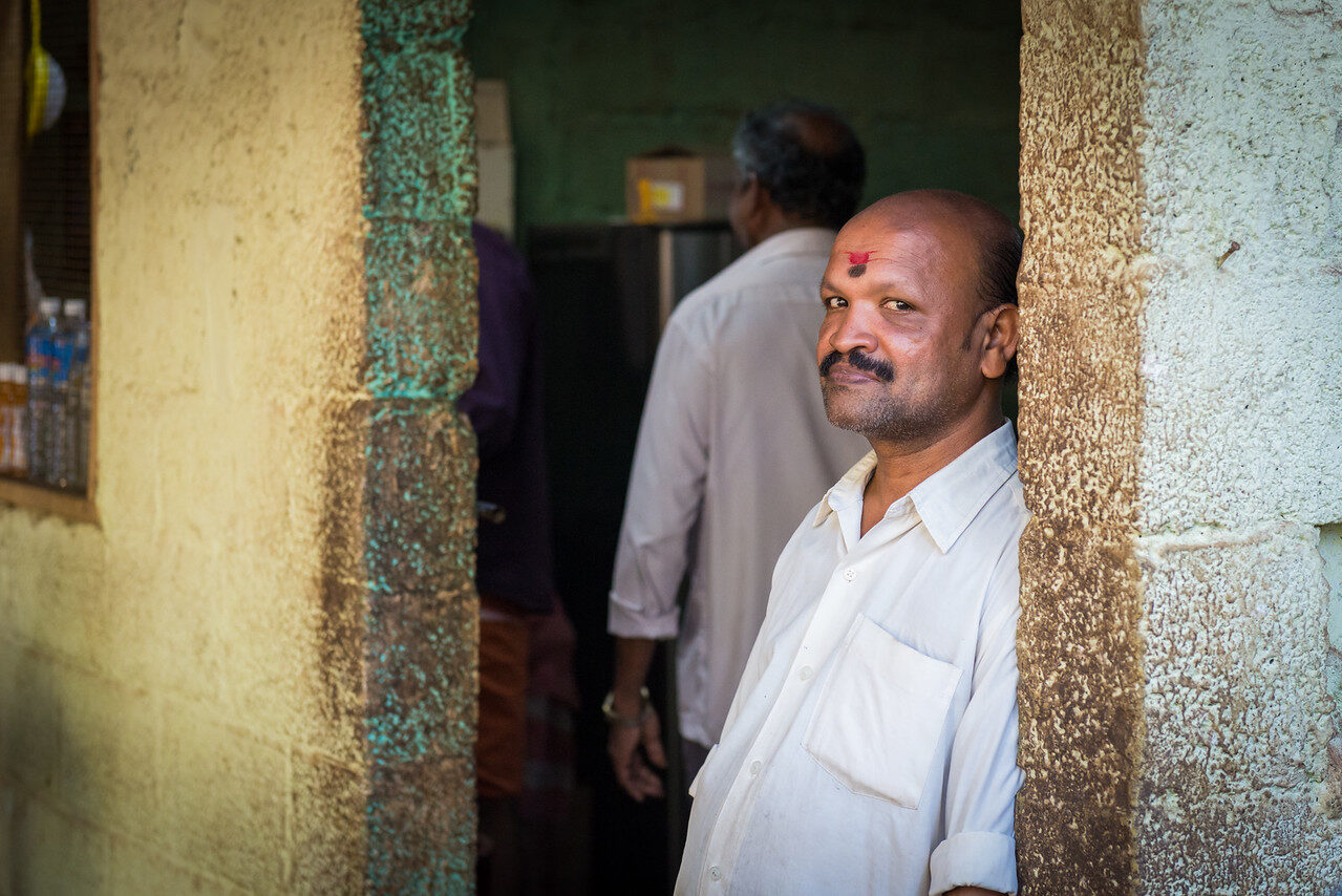 Man in a white shirt leans against a weathered doorway and looks toward the camera, with another man blurred inside behind him.