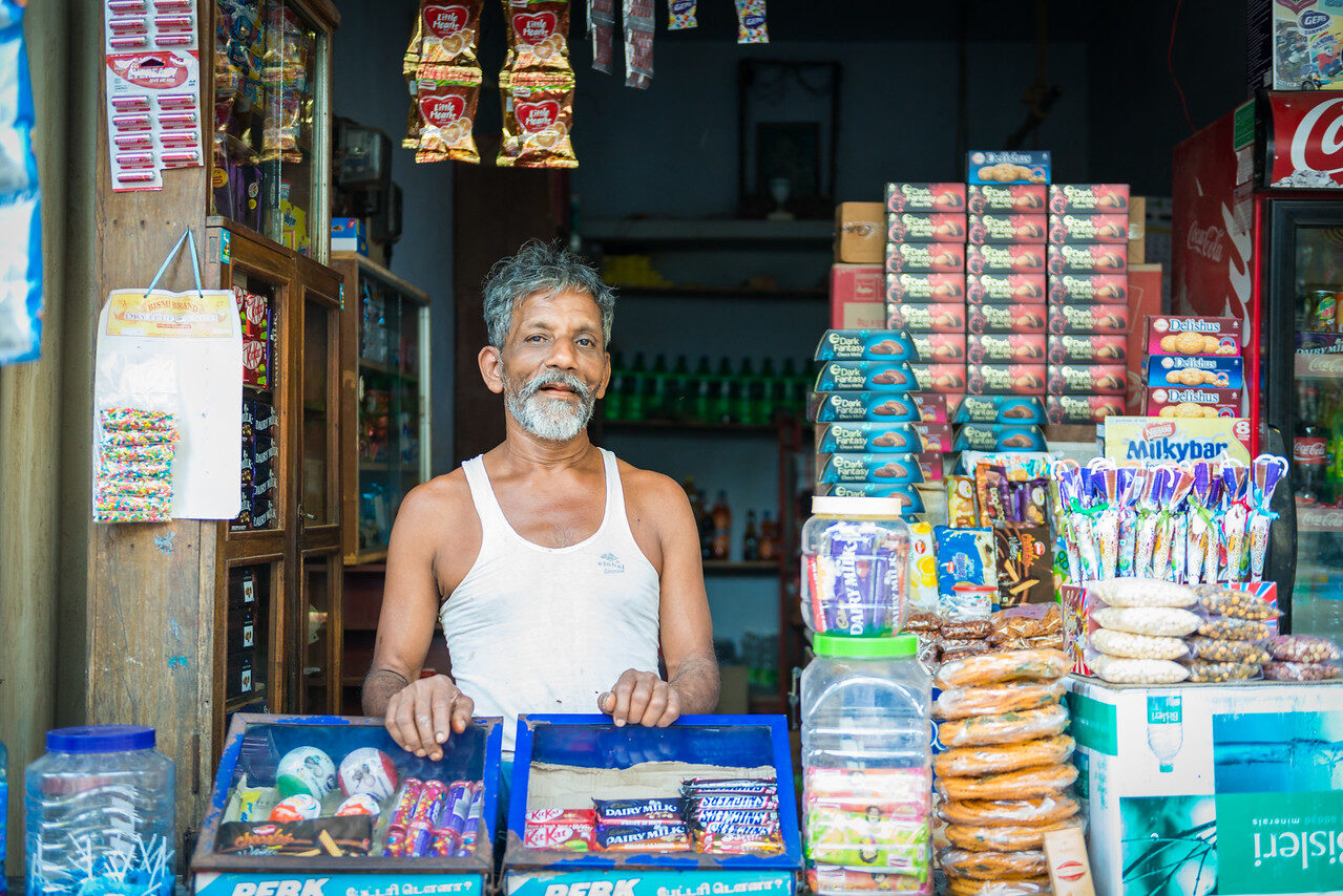 Gray-haired shopkeeper stands behind a counter crowded with snacks and candy in a small store.