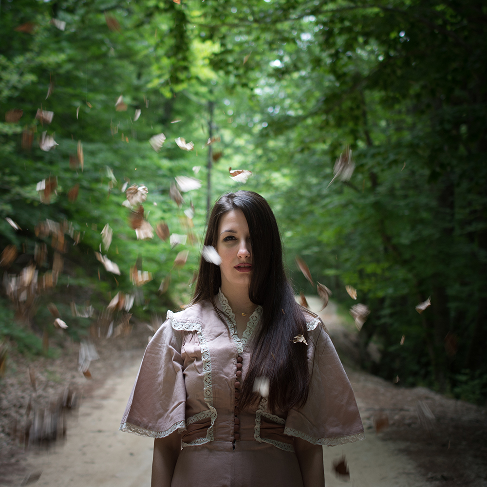 A woman stands on a wooded path as leaves fall around her.