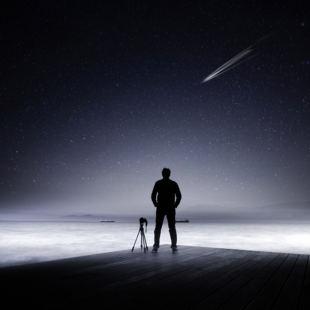 A person and a camera tripod stand on a deck by the water beneath a starry sky.