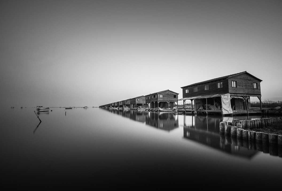 A row of wooden houses on stilts extends along still water in black and white.