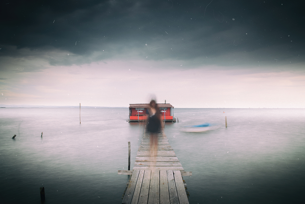 A blurred figure stands on a pier leading to a red house on the water.