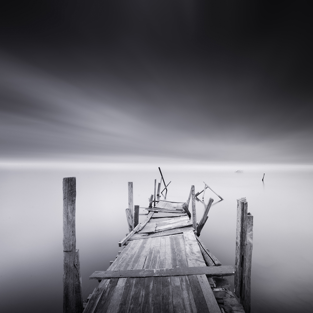 Black-and-white view down a broken wooden pier into still water.