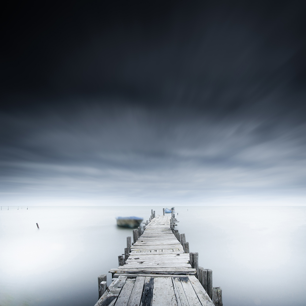 A weathered pier stretches into pale water beneath a dark clouded sky.