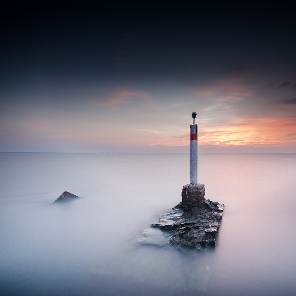 A striped post stands on rocks in smooth water at sunset.