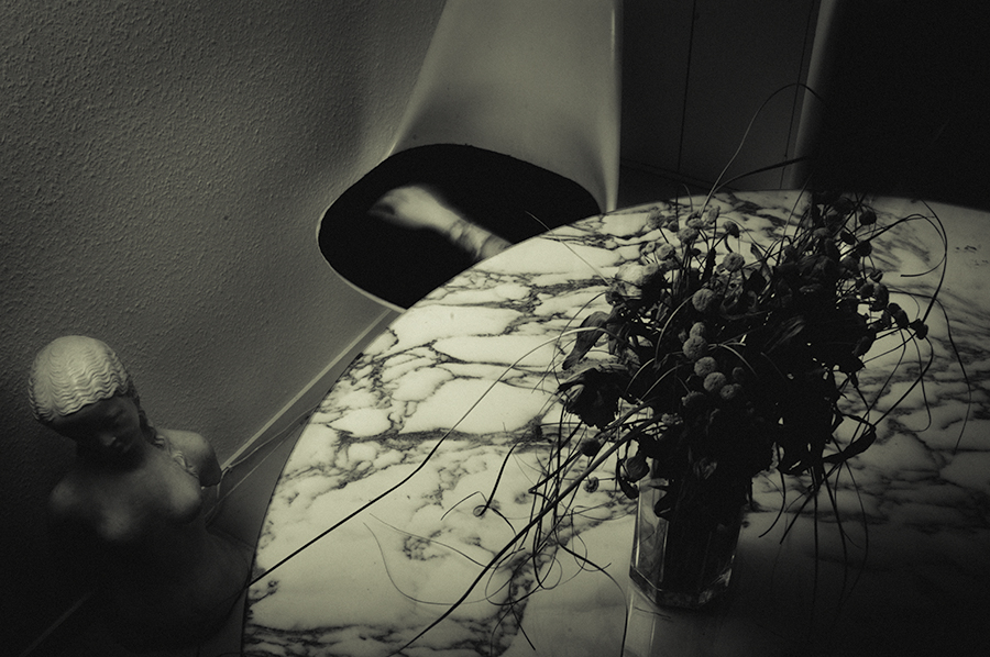Dim black-and-white room with a round table and flowers; a blurred figure is partly visible at left.