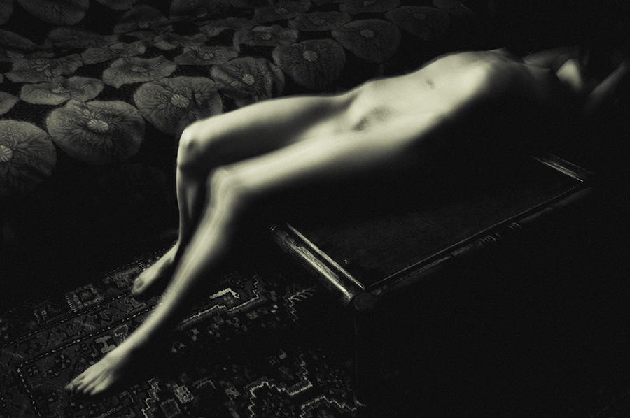 Blurred nude figure reclining on a patterned sofa beside a low wooden table.
